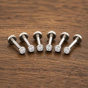 Puede incluir: Seis pendientes de labret de metal plateado, cada uno con una piedra preciosa redonda, transparente y facetada en un engaste de bisel al final de un poste recto con un disco plano en la parte posterior, dispuestos en fila sobre una superficie marrón.