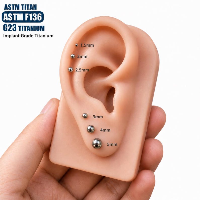 May include: ASTM F136 G23 implant grade titanium ball studs displayed on a beige ear model. The set includes six silver-coloured spherical piercings in graduated sizes labelled 1.5mm, 2mm, 2.5mm, 3mm, 4mm, and 5mm, showing the diameter of each piece.