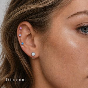 Könnte beinhalten: Ein Set aus vier Titan-Ohrsteckern mit runden Cabochon-Steinen in Pink, Hellblau, leuchtendem Blau und Weiß. Die Ohrringe sind mit den Größen 3mm, 3mm, 4mm und 5mm beschriftet und zeigen eine Größenabstufung entlang des Ohrknorpels.