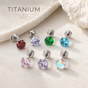 Könnte beinhalten: Sieben Titan-Ohrstecker mit rund geschliffenen Edelsteinen in Rot, Hellviolett, Grün, Blau, Rosa, Lavendel und schillerndem Weiß. Jeder Ohrstecker hat einen Metallstift und einen flachen runden Verschluss, mit dem Wort TITANIUM darüber.