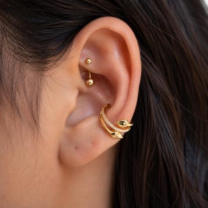 May include: A gold-toned ear cuff featuring a double-band design with snake-shaped ends and small clear stones embedded along the bands. A small gold-toned ball stud earring is positioned in the upper cartilage area of the ear.