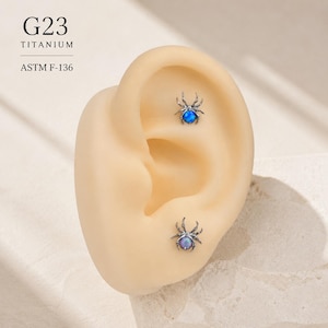 Könnte beinhalten: Zwei spinnenförmige Ohrringe aus G23-Titan, ASTM F-136. Jeder Ohrring hat ein dunkles Metallic-Finish und einen runden, schillernden Stein, der in der Mitte des Spinnenkörpers sitzt. Ein Stein ist blau, der andere hat einen hellvioletten Farbton.