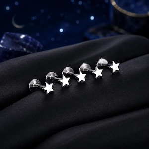 May include: Five silver-toned labret studs, each featuring a flat circular base and a five-pointed star-shaped top. The jewelry pieces are arranged in a row, showcasing a polished metallic finish and a simple, clean design for body piercings.