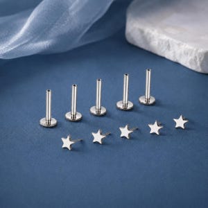 Może przedstawiać: Zestaw pięciu metalowych kolczyków typu labret w kolorze srebrnym, z których każdy posiada płaską okrągłą podstawę, prosty słupek i odpinaną końcówkę w kształcie gwiazdy. Elementy są ułożone w dwóch rzędach, z pionowymi słupkami i gwiazdkami.