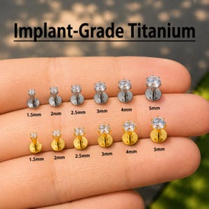 May include: A collection of implant-grade titanium labret studs with clear round-cut gemstones. The studs are arranged in two rows, one silver-toned and one gold-toned, with sizes labeled from 1.5mm to 5mm beneath each piece for clear identification.