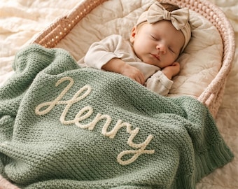 May include: A sage green knitted baby blanket featuring the name Henry embroidered in cream-colored yarn. The blanket is draped over a sleeping infant wearing a cream-colored long-sleeve shirt and a matching cream-colored headband with a bow.
