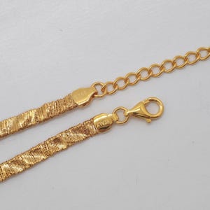 Op de afbeelding: Een goudkleurige ketting met een plat, getextureerd, geweven schakelontwerp. De ketting is voorzien van een karabijnsluiting en een verstelbare verlengketting met ronde schakels. Het metalen label is gestempeld met de tekst 925 en ITALY.