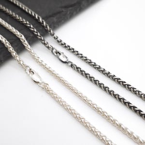 May include: Two wheat chain necklaces, one in a bright silver tone and one in a dark oxidized gunmetal gray tone. Each chain features a woven, braided link pattern and is secured with a polished lobster claw clasp at the end of the metal chain.
