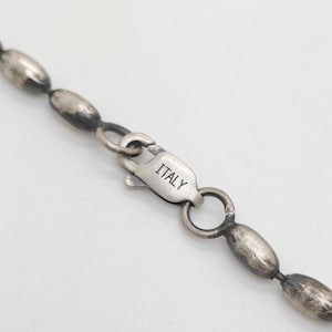 Pode incluir: Um colar de corrente em tom prateado com contas ovais alongadas e um acabamento escuro e oxidado. O colar inclui um fecho tipo lagosta com a palavra ITALY claramente estampada na superfície metálica do fecho.