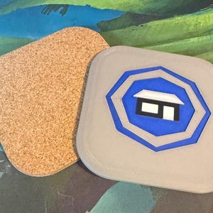 Op de afbeelding: Twee vierkante onderzetters met afgeronde hoeken. Eén onderzetter heeft een bovenkant van lichtbruin kurk. De andere is lichtgrijs met een centraal blauw achthoekig ontwerp met een wit-zwarte afbeelding van een klein gebouw met dak.