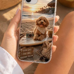 May include: A clear smartphone case featuring a printed beach scene with a golden doodle dog wearing a bandana on a surfboard, a vintage camera, a woven bag, and sunglasses. The text reads: GOOD DAYS, TAN LINES, WAGGING TAILS with a heart icon.