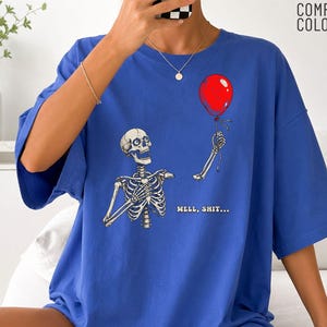 Peut inclure: Un t-shirt bleu roi avec une illustration de squelette tenant un ballon rouge et le texte "WELL, SHIT..." imprimé en jaune. Le vêtement présente des manches courtes et un col rond, avec la marque "COMFORT COLORS" visible dans le coin supérieur.