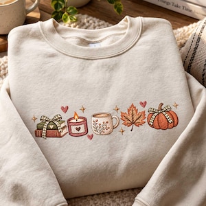 Puede incluir: Una sudadera de cuello redondo color crema con un diseño bordado en el pecho. El bordado muestra una pila de libros con un lazo a cuadros, una vela encendida, una taza con detalles florales, una hoja de arce y una calabaza con un lazo.