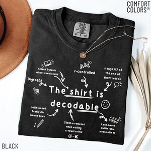 Op de afbeelding: Een zwart t-shirt met korte mouwen en witte grafische tekst die The shirt is decodable leest, omringd door educatieve taalkundige notities zoals Closed Syllable, r-controlled, Digraphs en Latijnse achtervoegsels, met een kleine gouden ketting.