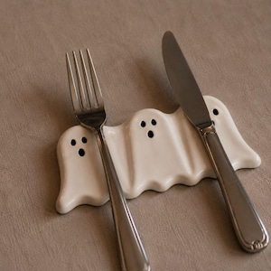 May include: A white ceramic utensil rest shaped like three ghosts with black painted eyes and mouths. The piece features three distinct indentations designed to hold cutlery, with a silver metal fork and a silver metal knife resting on top.