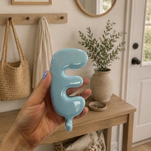 May include: A glossy, light blue, three-dimensional wall hook shaped like the capital letter E. The hook features a smooth, rounded, balloon-like aesthetic with a curved top and a small, protruding hook at the bottom for hanging items.