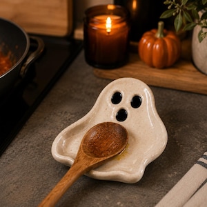 May include: A cream-colored ceramic spoon rest shaped like a ghost with three black oval cutouts for eyes and a mouth. The surface has a speckled texture. A wooden spoon with a rounded bowl rests inside the ghost-shaped ceramic dish.