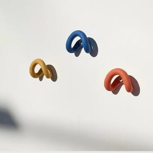 May include: Three wall-mounted hooks in a curved, loop-like shape. One hook is blue, one is yellow, and one is orange. Each hook features a smooth, matte finish and is attached to the wall with a small, circular metal base, casting a distinct shadow.