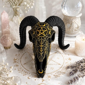 May include: A black ram skull sculpture featuring intricate gold filigree patterns, a prominent gold pentagram on the forehead, and a small gold crescent moon at the nasal cavity. The skull has large, curved, textured black horns on each side.