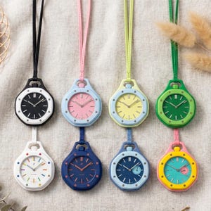 Könnte beinhalten: Acht Anhängeruhren mit Umhängebändern in verschiedenen Farben wie Schwarz, Pink, Limettengrün, Waldgrün, Weiß, Marineblau, Hellblau und Gelb. Jede Uhr verfügt über ein strukturiertes Zifferblatt, silberne Zeiger und ein rundes Gehäuse mit Öse.
