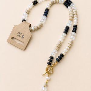 May include: A necklace featuring a mix of white pearls, black and beige disc beads, and clear faceted beads, finished with a gold-tone toggle clasp. The necklace is attached to a brown cardboard display tag with the text $20 written on it.
