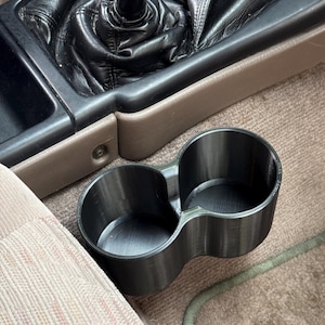 May include: A black, double-cup holder, likely 3D-printed, sits on a car's carpeted floor. The holder is positioned near the gear shift and a car seat. The interior appears to be tan and black.