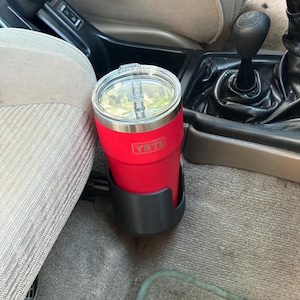May include: A bright red YETI tumbler with a clear lid and a straw, positioned securely within a black cup holder inside a vehicle. The tumbler features the YETI brand logo embossed on the side, and the clear lid allows a view of the straw inside.