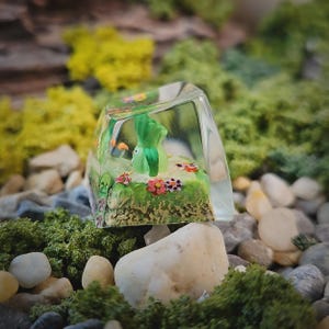 May include: A clear resin mechanical keyboard keycap featuring an encapsulated miniature scene with a small green creature, textured green ground, and tiny colorful flowers. The keycap has a smooth, transparent, angled top surface.