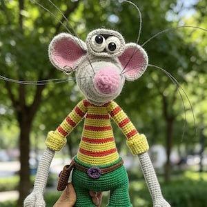 May include: A crocheted mouse doll with a grey head, pink nose, and large ears. It wears a yellow and red striped shirt, green shorts with a brown belt, and blue shoes. The doll has long grey arms, striped yellow and blue leg warmers, and long whiskers.