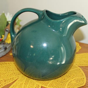 May include: A dark green ceramic pitcher with a spherical body, a curved handle, and a wide pouring spout. The surface of the pitcher has a smooth, glossy finish that reflects light, highlighting its rounded shape and solid, deep green color.