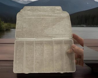 May include: A clear, rectangular plastic storage container featuring a hinged lid and six vertical interior compartments. The organizer is constructed from a translucent material, allowing for visibility of the contents within each section.