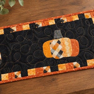 May include: A rectangular quilted table runner featuring a black center panel with swirling stitch patterns and two fabric pumpkins. The border consists of alternating orange, black, and white checkered squares with an orange outer edge trim.