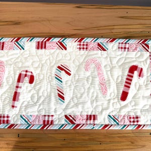 Puede incluir: Un camino de mesa acolchado rectangular con fondo blanco y un patrón de costura en espiral. El centro muestra cinco aplicaciones de bastones de caramelo con patrones en rojo, rosa y turquesa. El borde consiste en cuadrados alternos de tela a cuadros y rayas.