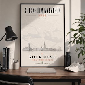 Op de afbeelding: Een ingelijste poster voor de Stockholm Marathon 2026, met een kaart en een illustratie van de skyline van de stad. De poster bevat de tekst "YOUR NAME" en "FINISH TIME 03:42:15". De poster staat op een houten bureau met een laptop, hardloopschoenen en een waterfles.