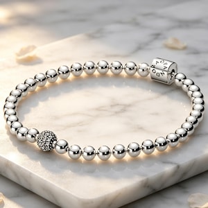 May include: A silver beaded bracelet featuring a series of polished metallic spheres, a single textured bead encrusted with small clear stones, and a cylindrical clasp engraved with the word PANDORA in a clean, circular arrangement.