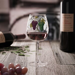 May include: A clear glass wine goblet featuring a painted design of purple and green grape clusters with green leaves. The glass is filled with red wine and stands on a slender stem with a circular base, showcasing detailed botanical illustrations.