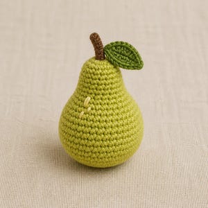 May include: A crocheted pear in a light green yarn with a textured surface. The fruit features a short brown stem at the top and a single dark green crocheted leaf attached to the side. A few small yellow stitches are visible on the side of the pear.