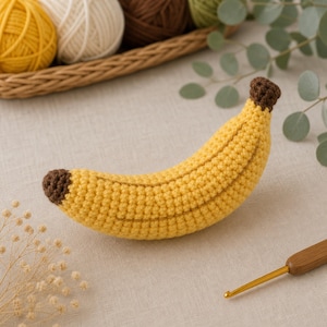 May include: A handmade crochet banana toy featuring a bright yellow curved body with textured stitches and dark brown tips at both ends. The banana is crafted with a visible yarn construction, showcasing a detailed, soft, and rounded fruit shape.