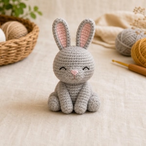 Peut inclure: Un petit lapin en crochet fait main avec un corps gris clair, de longues oreilles aux détails intérieurs rose pâle et un petit nez brodé en rose. Le lapin a une tête arrondie, une posture assise et des yeux fermés brodés en forme d'arc.