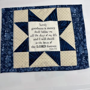 Può includere: Un arazzo rettangolare in tessuto con un motivo a stella centrale in tessuto blu navy e crema a pois. Il quadrato centrale mostra il testo: Surely goodness & mercy shall follow me all the days of my life and I will dwell in the house of the LORD forever. Psalm 23:6.