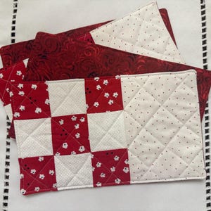 May include: A set of rectangular quilted placemats featuring a patchwork design of red and white floral fabric squares alongside a solid white section with small black dots, all stitched with a diamond-shaped quilting pattern on the white areas.