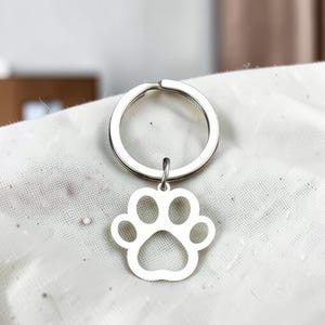 May include: A silver-toned metal keychain featuring a circular split ring attached to a flat, cutout charm in the shape of a paw print. The charm has a smooth, polished finish and is centered below the ring, showcasing a simple, open-frame design.