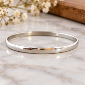 Pode incluir: Uma pulseira tipo bracelete de metal prateado polido com uma superfície lisa e arredondada e um design circular contínuo. A pulseira apresenta um acabamento reflexivo que capta a luz, criando uma estética limpa e simples para o pulso.