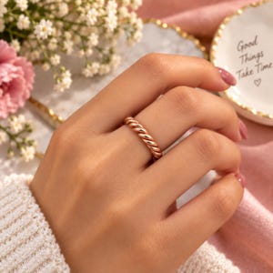 May include: A rose gold ring featuring a twisted rope design worn on a finger. The band has a polished metallic finish with a continuous spiral pattern that wraps around the entire circumference of the ring, creating a textured appearance.
