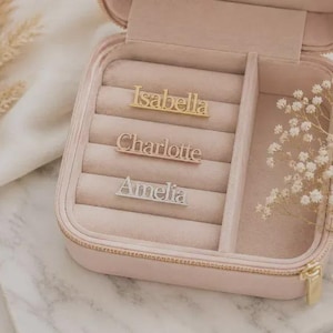 May include: A light pink square jewellery box with a velvet interior containing three horizontal metal name bars. The top bar is gold with the name Isabella, the middle is rose gold with Charlotte, and the bottom is silver with the name Amelia.