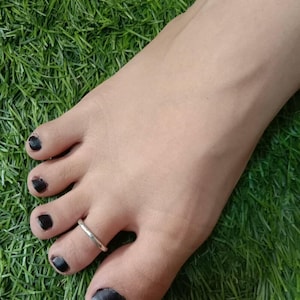 Puede incluir: Un anillo para el dedo del pie de metal plateado, liso y sencillo, colocado en el segundo dedo. El anillo presenta un diseño de banda pulida y circular. Las uñas de los pies están pintadas con un esmalte de color negro sólido y opaco.