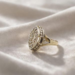 Op de afbeelding: Een goudkleurige metalen ring met een ovaalvormig medaillon voorzien van ingewikkeld filigraan krulwerk en een kralenrand. Het medaillon is bevestigd aan een gladde ringband met een gespleten schouder en heeft een scharniermechanisme aan de zijkant.