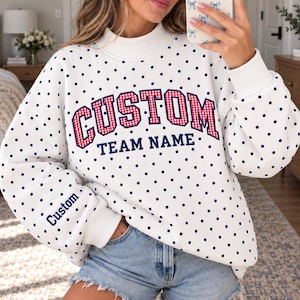 May include: A white long-sleeve sweatshirt with a navy blue polka dot pattern, featuring the text CUSTOM TEAM NAME in red and white gingham lettering on the chest and the word Custom embroidered in navy blue on the sleeve cuff.