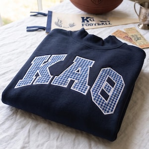 Könnte beinhalten: Ein marineblaues Sweatshirt mit Rundhalsausschnitt und den griechischen Buchstaben KAO, die in einem blau-weißen Vichy-Muster mit weißer Stickkante appliziert sind. Die Buchstaben sind mittig auf der Brust des auf einer weißen Fläche liegenden Kleidungsstücks.