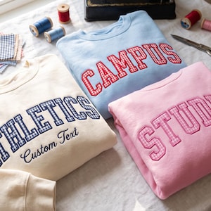 May include: Three crewneck sweatshirts in cream, light blue, and pink. The cream sweatshirt features the word ATHLETICS in blue gingham and the text Custom Text. The blue sweatshirt says CAMPUS in red gingham, and the pink one says STUDIOS in pink gingham.